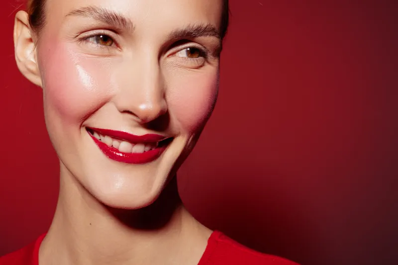 Close-up of smiling woman with red lipstick and glowing skin on red background. Concept of beauty, makeup, skincare, and happiness with natural light and vibrant colors.