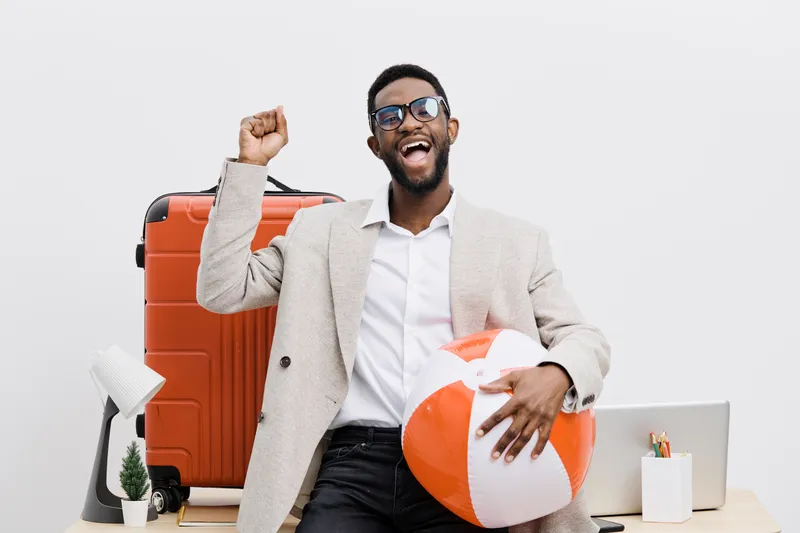 Joyful smiling man with dark skin wearing glasses, light-colored blazer, holding a beach ball, surrounded by travel accessories including a large orange suitcase, laptop, and plant in a studio setting