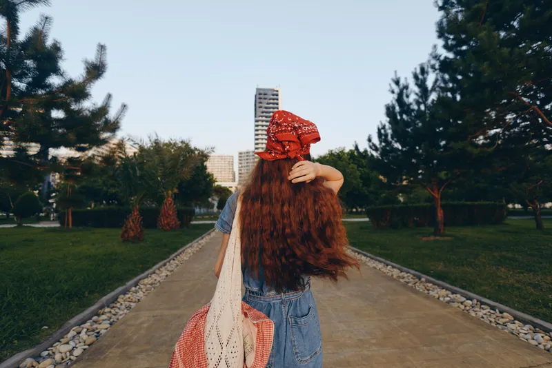 lifestyle woman in modern boho-western denim street style walking outdoors with long hair and red bandana, film color tone, casual summer outfit in urban park setting