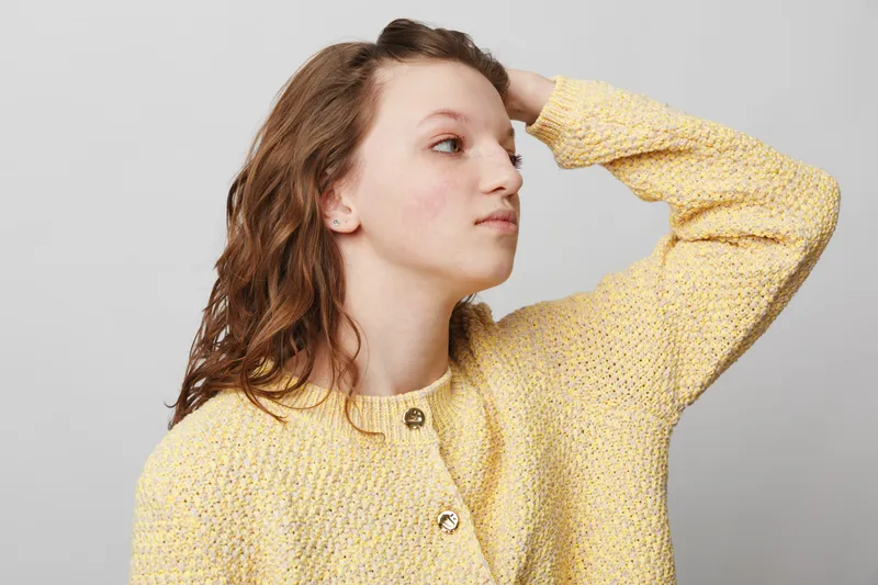 Young woman in yellow knitted sweater looks sideways with hand on head. Studio portrait on grey background. Concept of casual fashion, thoughtful mood, and natural beauty.