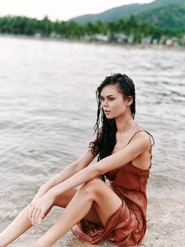 thoughtful woman sitting on the beach wearing a brown dress with wet hair and calm sea background under soft daylight