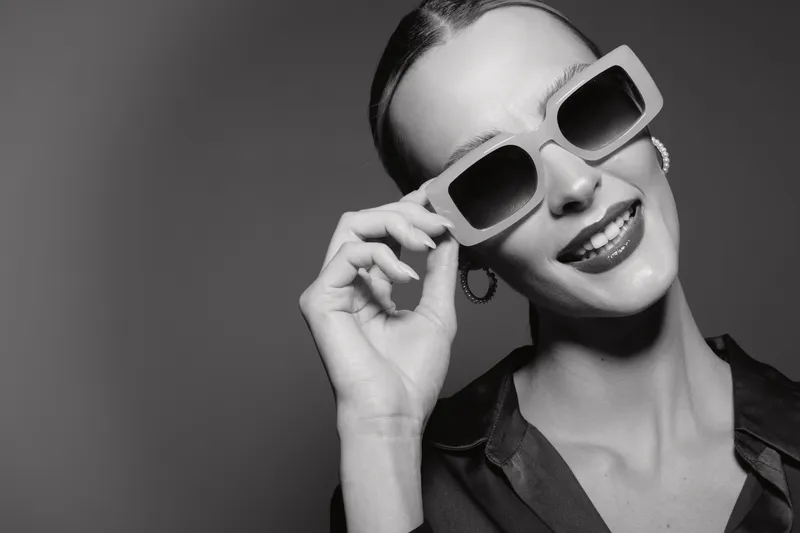 Elegant young white woman with pale skin and slicked hair wearing sunglasses smiles confidently on a dark background. Concept of fashion, style, beauty, and confidence.