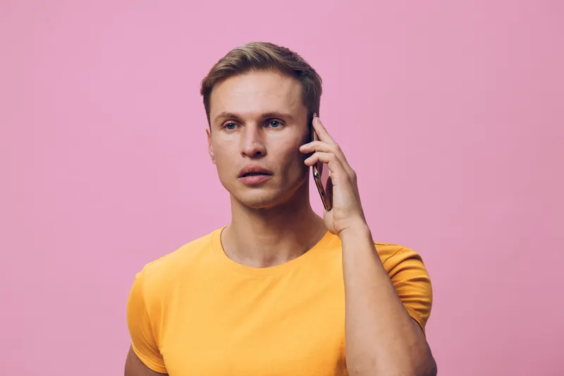 Full body portrait of a young man with short hair wearing a yellow t-shirt talking on mobile phone against pink background, indoor studio shot, casual style, lifestyle concept, communication technolog