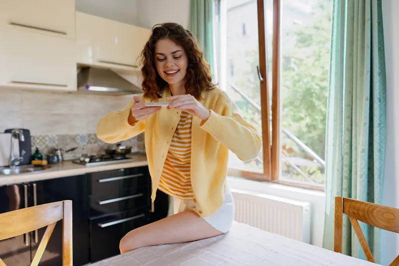 Young woman with curly hair sits on a kitchen table wearing a yellow cardigan and striped top, smiling at a phone in a bright home setting with soft natural light from a large window