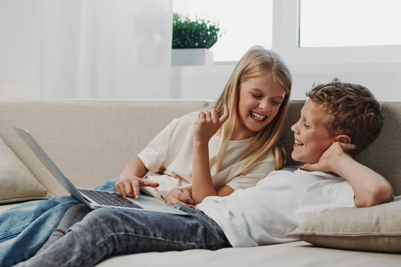 Happy siblings relax on couch with laptop, smiling and talking in bright living room. Concept of family bonding, leisure, and technology use at home.