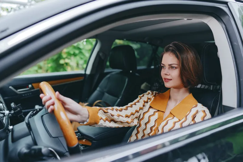 A woman sits behind the wheel of a car, steady hands on the steering, looking forward with calm concentration during daytime drive.