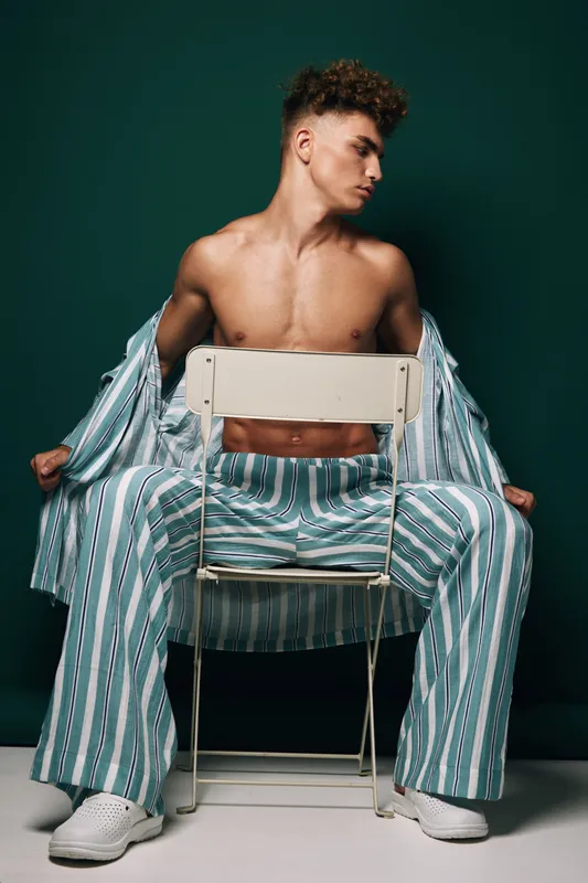 Young shirtless man in striped pajama pants poses sitting backward on chair with open shirt for fashion portrait against solid dark background in studio. Casual striped outfit and wife beater.