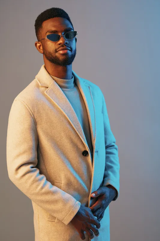 Stylish African man in beige coat and sunglasses, showcasing confidence against a blue backdrop, representative of modern fashion trends and sophistication