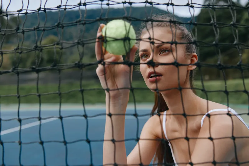 Young athletic woman peering through tennis net while holding tennis ball, expressing focus and determination on a sunny day outdoors