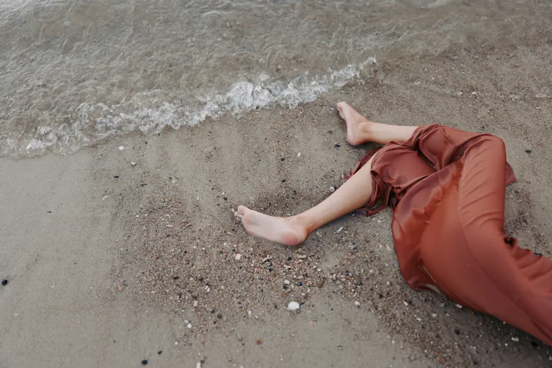 A single leg and rust red pants rest on damp sand beside a calm shoreline, with soft water ripples and light reflecting on the surface at the foamy edge.