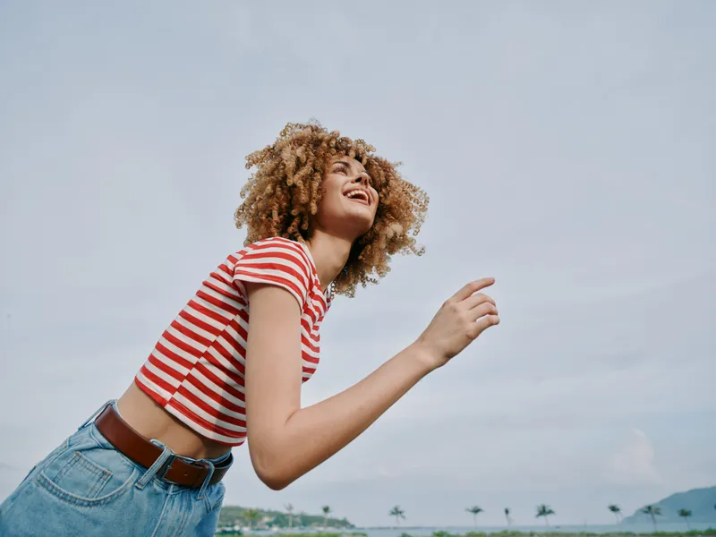 Young woman with curly hair in a red striped shirt and jeans stands in an open outdoor setting, smiling warmly as she leans forward in a carefree pose.