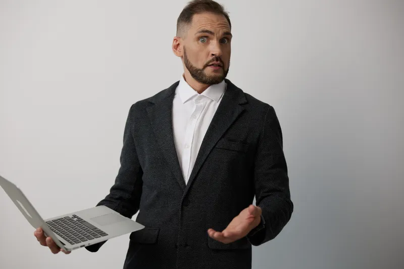 A professional man in a dark patterned suit holds a sleek laptop against an isolated colored background, projecting a confident, curious mood and a decisive, ready-to-act gesture for business