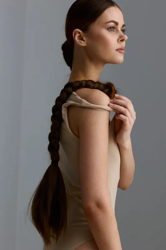 Young woman with long braided hair, wearing a beige sleeveless top, posing thoughtfully in soft natural light against a minimalist gray background.