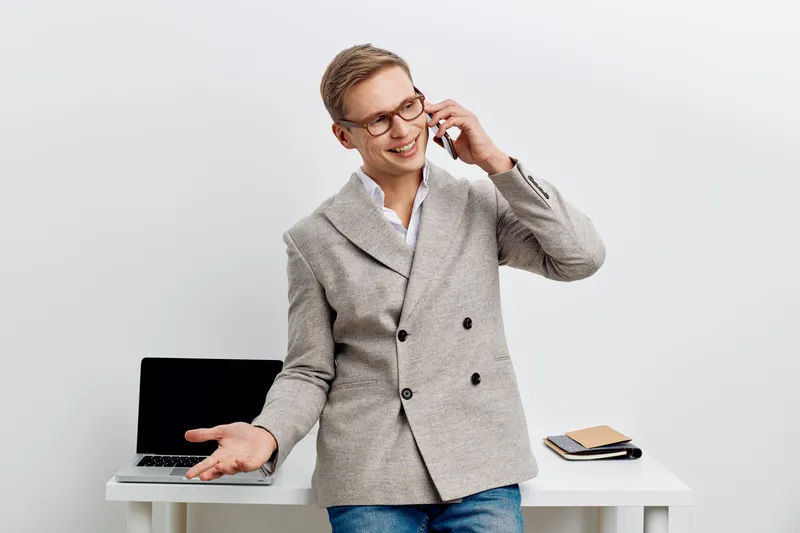 smiling man talking phone casual jacket jeans standing desk laptop digital nomad remote work communication concept