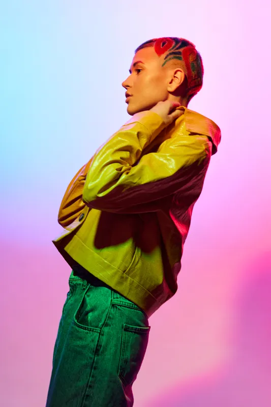 Stylish woman with colorful shaved hair wearing a yellow jacket and blue jeans posing confidently against a vibrant pink and purple gradient background.