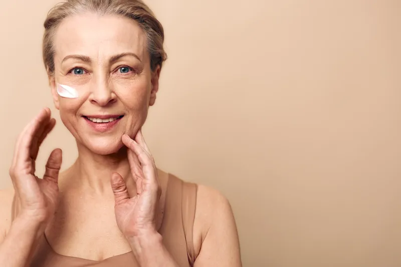 Smiling mature woman with natural skin applies moisturizing cream on her face. Beige background and soft lighting create a warm atmosphere. Concept of skincare and anti aging.