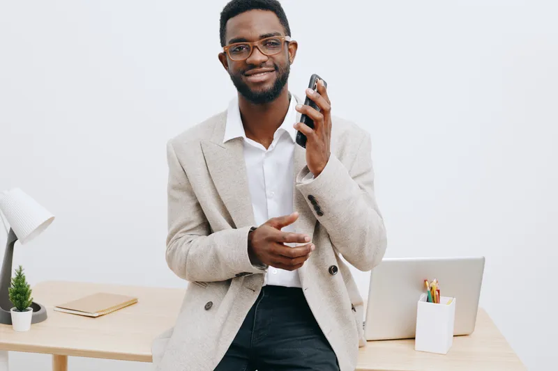 Smiling man with glasses holding smartphone in office setting, dressed in beige blazer and white shirt, standing near desk with minimal decor, modern business lifestyle concept.