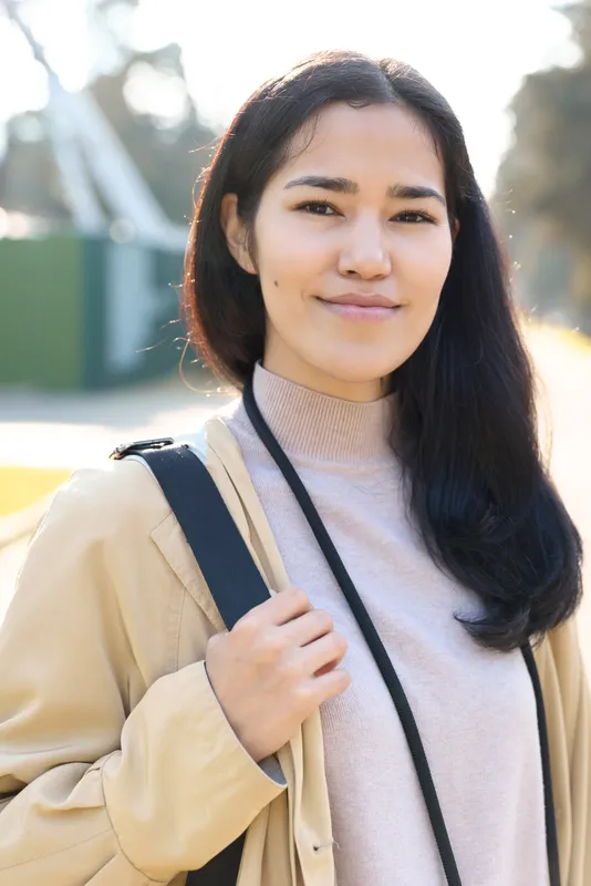 Candid street style, a confident woman with a backpack, beige coat and soft sweater, standing outdoors in morning light, calm smile, urban lifestyle and travel vibe.