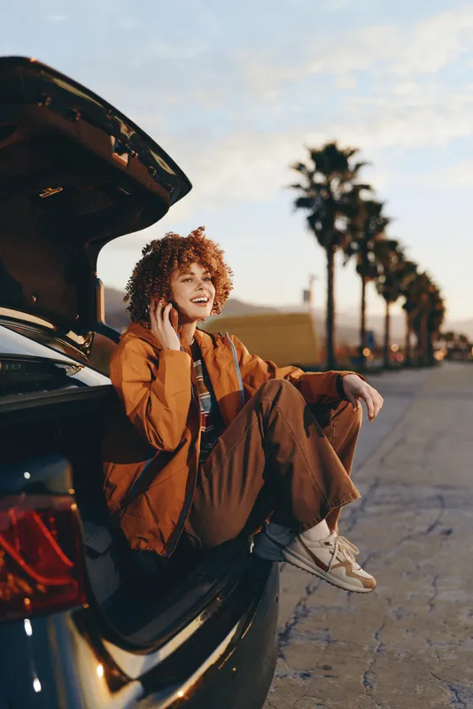 Woman lifestyle outdoors by car trunk, smiling with curly hair, wearing warm jacket, relaxed travel moment, sunny road daytime, parked vehicle, casual stance, natural light