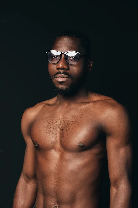African man with dark skin and glasses posing shirtless in creative conceptual visual style against black background. Portrait of confident male model with natural expression and strong body.