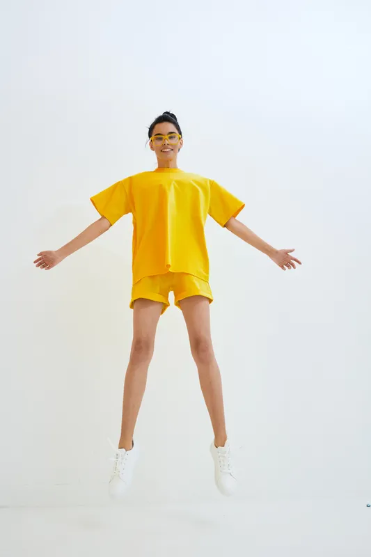 Woman jumping in yellow casual outfit against white background, wearing sunglasses and sneakers, expressing joy and energy with open arms in bright minimalistic setting.