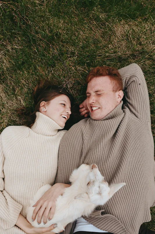 A couple lying on grass in a cozy outdoors scene, smiling at each other with a fluffy cat between them, capturing a lifestyle moment of warmth and affection.