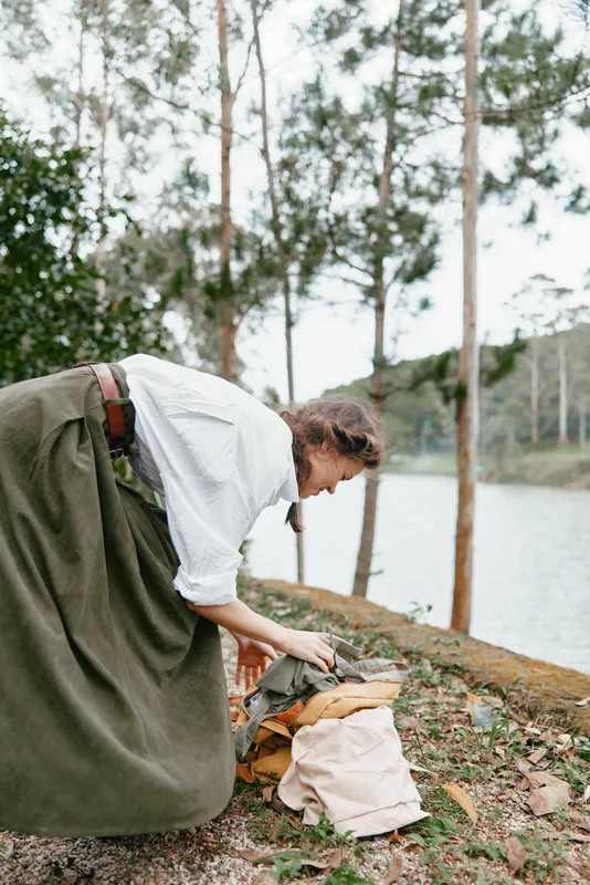 Woman in traditional attire organizing materials by a serene lake surrounded by trees, evoking a sense of creativity and exploration in nature