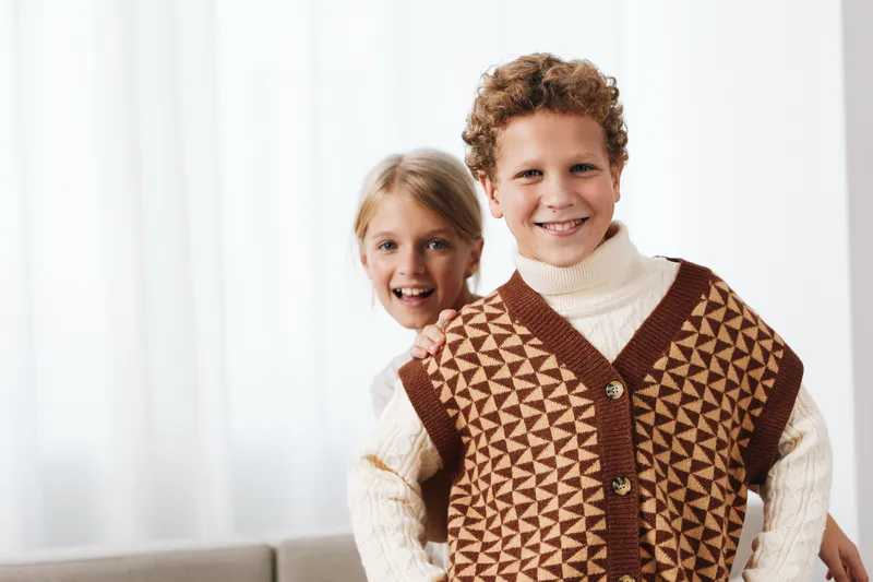 Authentic portrait of children in a cozy home setting, wearing a knit vest and school inspired attire, capturing a playful, candid moment with warm light.