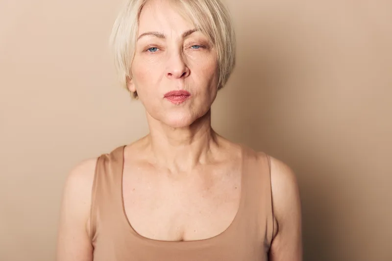 Portrait of a serious mature woman with short blonde hair wearing a beige tank top against a neutral background. Concept for aging, confidence, and natural beauty in minimal style.