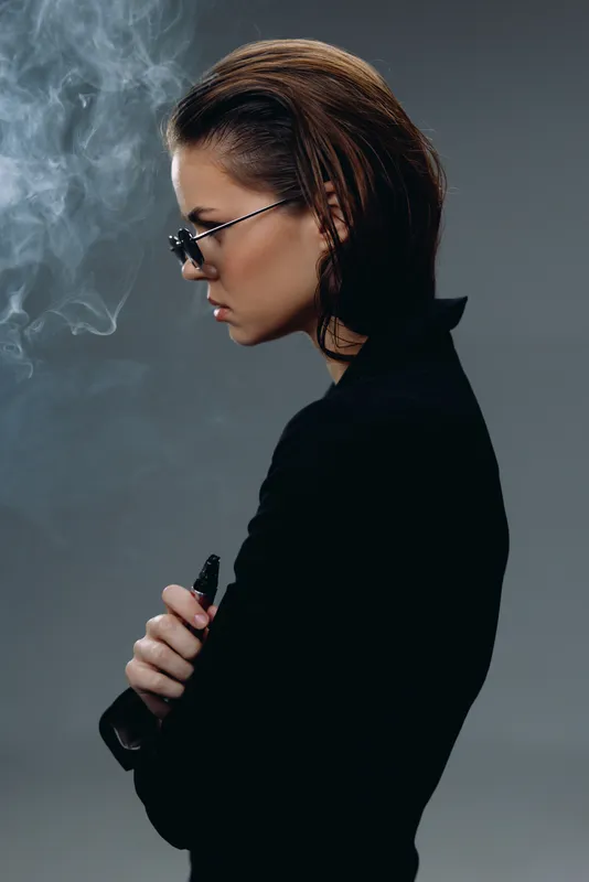 Profile of a young woman vaping with smoke against a dark background, wearing glasses and a black sweater.