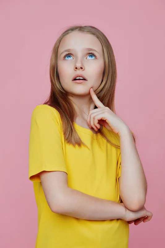 Young girl in a yellow shirt looks thoughtfully upwards against a pink background, expressing curiosity and contemplation