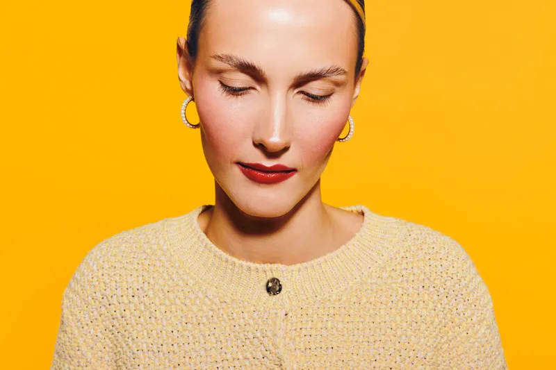Young woman with closed eyes and red lipstick wears yellow sweater and gold earrings on bright yellow background. Concept of calm, beauty, fashion, and minimalism.