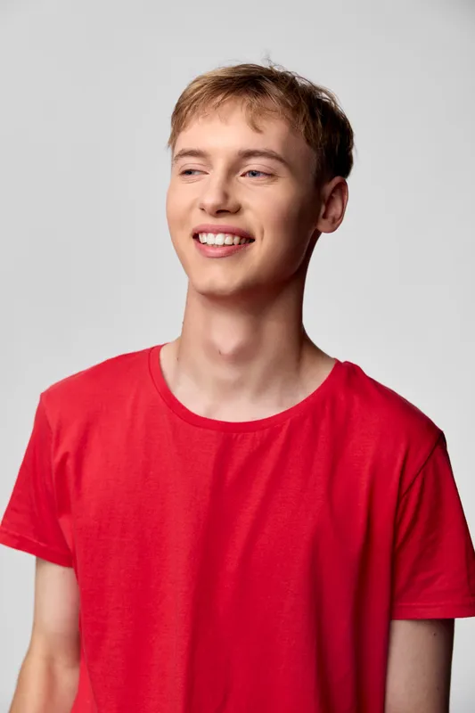 Smiling young man in a red shirt stands in a bright studio, conveying a friendly casual vibe ideal for lifestyle, fashion, or marketing campaigns focused on positivity and everyday youth.