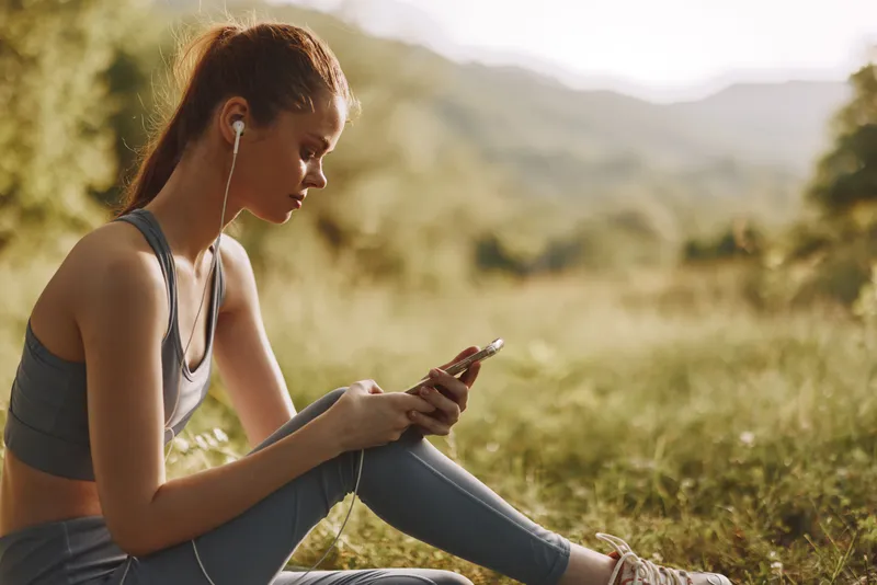 Young woman in sportswear sitting on grass, listening to music on her phone, enjoying nature in a serene outdoor setting, ideal for wellness and fitness themes