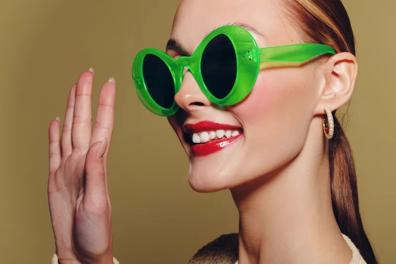 Smiling woman with green sunglasses and red lipstick waves hand. Close-up portrait on beige background. Concept of fashion, summer style, and positive mood in studio lighting.