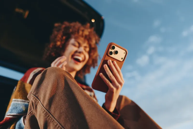 Woman with curly hair smiling in car holding smartphone for selfie, driving lifestyle moment, bright daylight through window, casual travel mood, candid snapshot summer outdoors