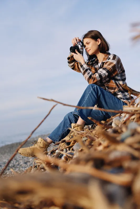 Photographer, woman perched on coastal rocks, capturing landscape with a camera, amid autumn leaves, ocean breeze and wide sky, outdoor nature scene