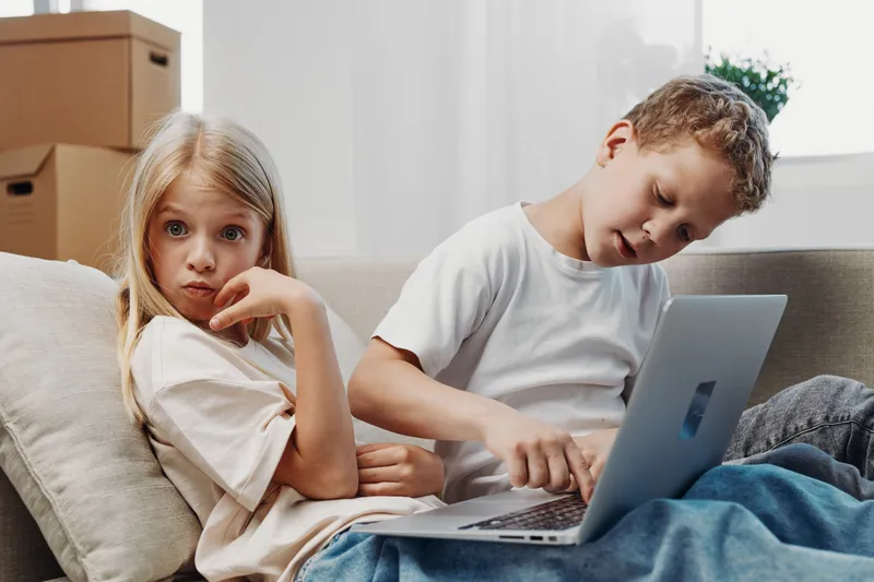 Two children relax on a couch with a laptop, a boy focused on the screen and a girl looking thoughtfully. Natural light illuminates the casual home environment and sibling interaction.