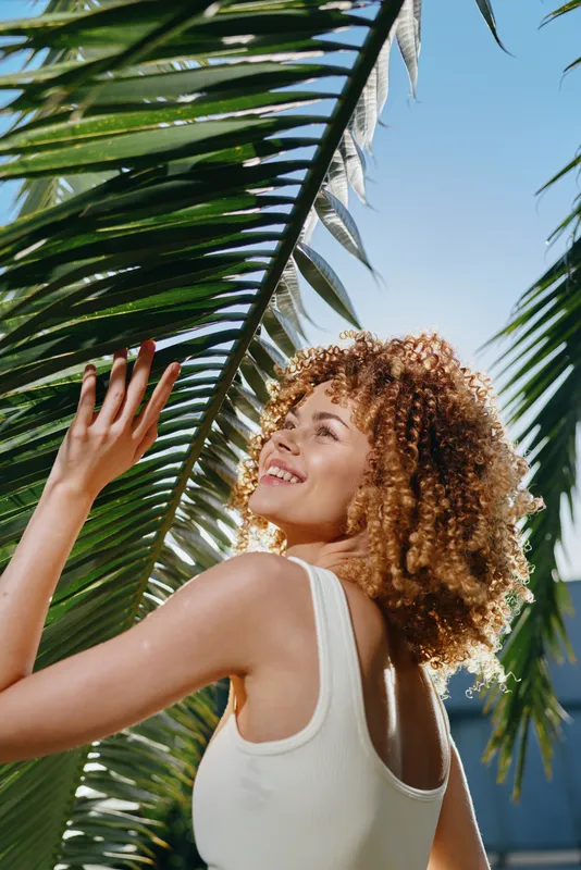 smiling woman with curly hair enjoying tropical sun, white sleeveless top, lush green palm leaves, bright blue sky, natural summer outdoor setting