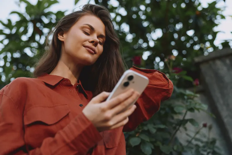Smiling woman in a rust orange shirt holds a smartphone outdoors, capturing a casual lifestyle moment. Natural greenery and soft light create a warm, relaxed mood