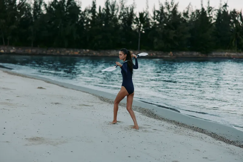 A lone woman walks the sandy shoreline with a surfboard over her shoulder, preparing for a seaside session as calm blue water and a forest backdrop create a serene scene.