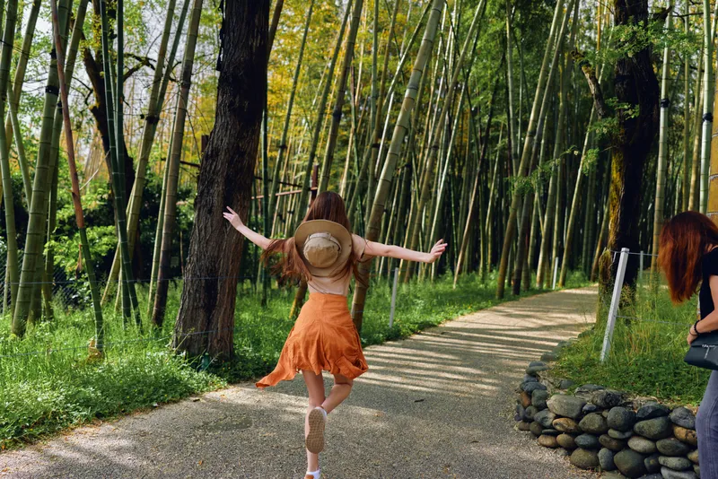 Woman in orange dress and straw hat walking joyfully on a pebble path through tall bamboo forest, arms outstretched, summer travel, carefree nature scene and outdoor exploration.