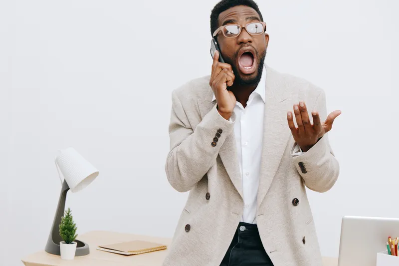 Young professional man with glasses in a beige blazer talking on mobile phone while showing confusion or frustration, standing next to a desk with a laptop, plant, and notebook on plain white backgrou