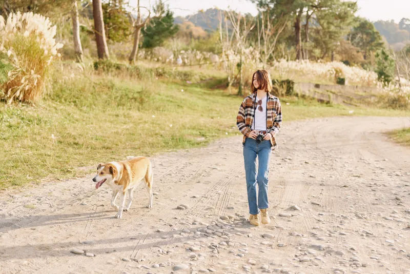 Woman walking a dog along a gravel path in outdoor countryside during sunny light, casual fashion visible in plaid shirt and jeans, capturing companionship on a peaceful nature walk