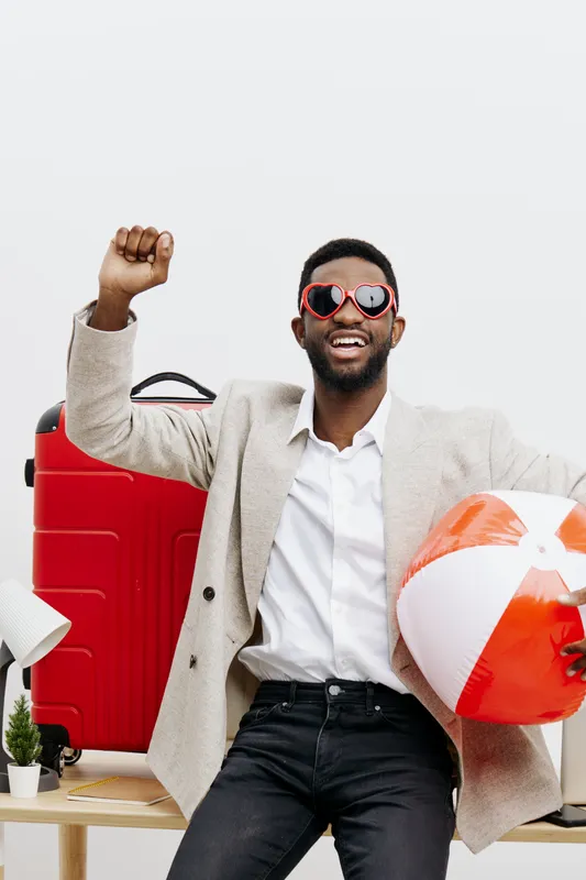 Happy cheerful man with dark skin wearing stylish sunglasses holding inflatable beach ball and red suitcase. Man in casual outfit smiling excitedly against white background. Summer holiday, travel con