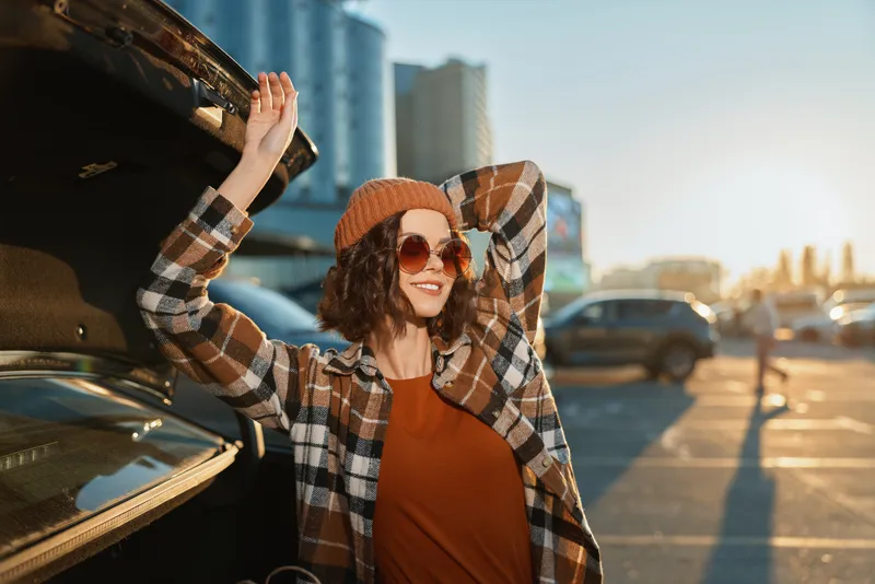 Woman with sunglasses and smile leaning on car trunk in urban parking at sunset, authenticity and candid lifestyle captured with golden hour glow, mindful living and emotional storytelling.