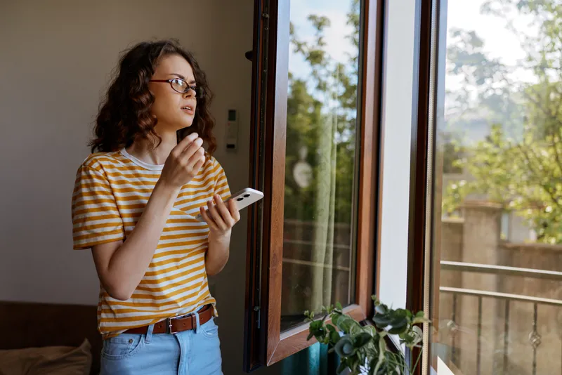 Casual woman at home looking out the window, wearing striped tee and jeans, holding a smartphone, natural sunlight, relaxed lifestyle moment, peaceful indoor scene