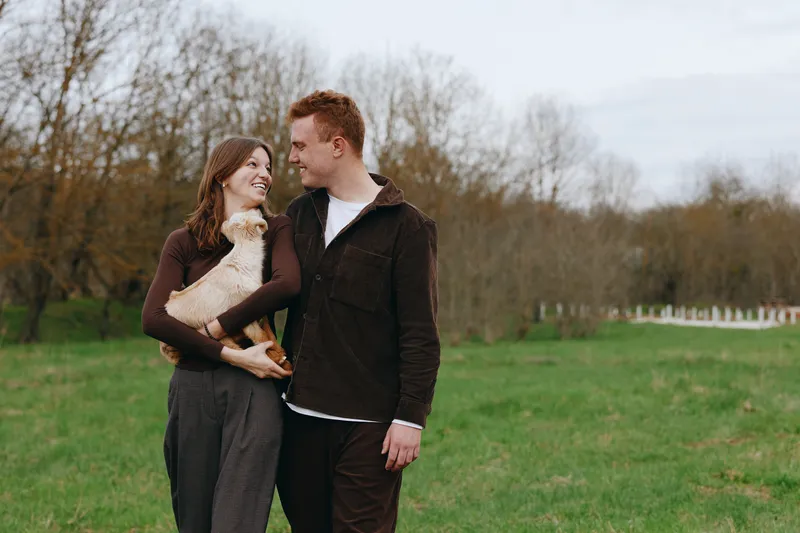 A couple outdoors with a goat kid in a grassy field, sharing a casual moment and lighthearted affection. Natural setting emphasizes lifestyle, nature, and gentle companionship.