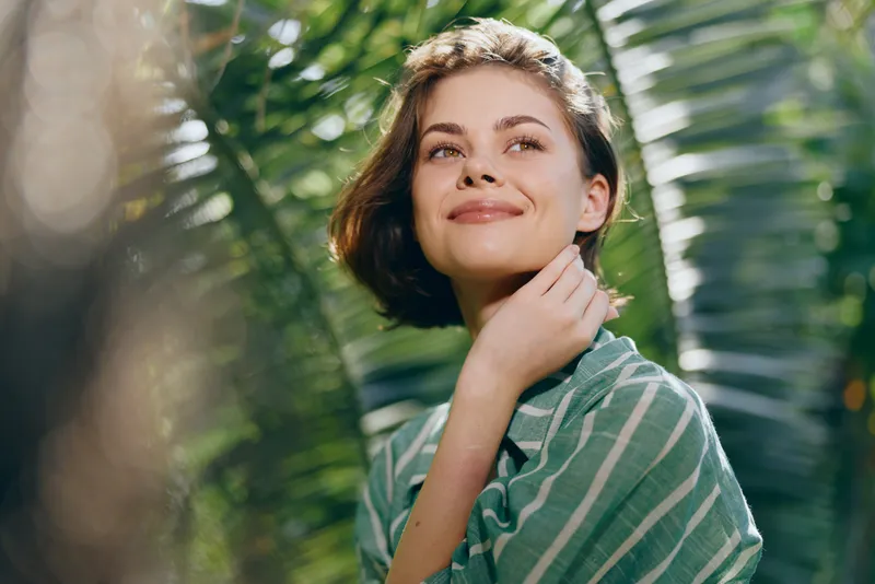 A woman smiles softly in a lush outdoor setting, posing relaxed and confident among greenery. This portrait captures natural light, warm atmosphere, and a serene, casual mood.