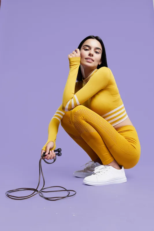 woman sport fitness in yellow activewear crouching with jump rope, dark hair, white sneakers, purple background, dynamic and energetic pose, healthy lifestyle concept